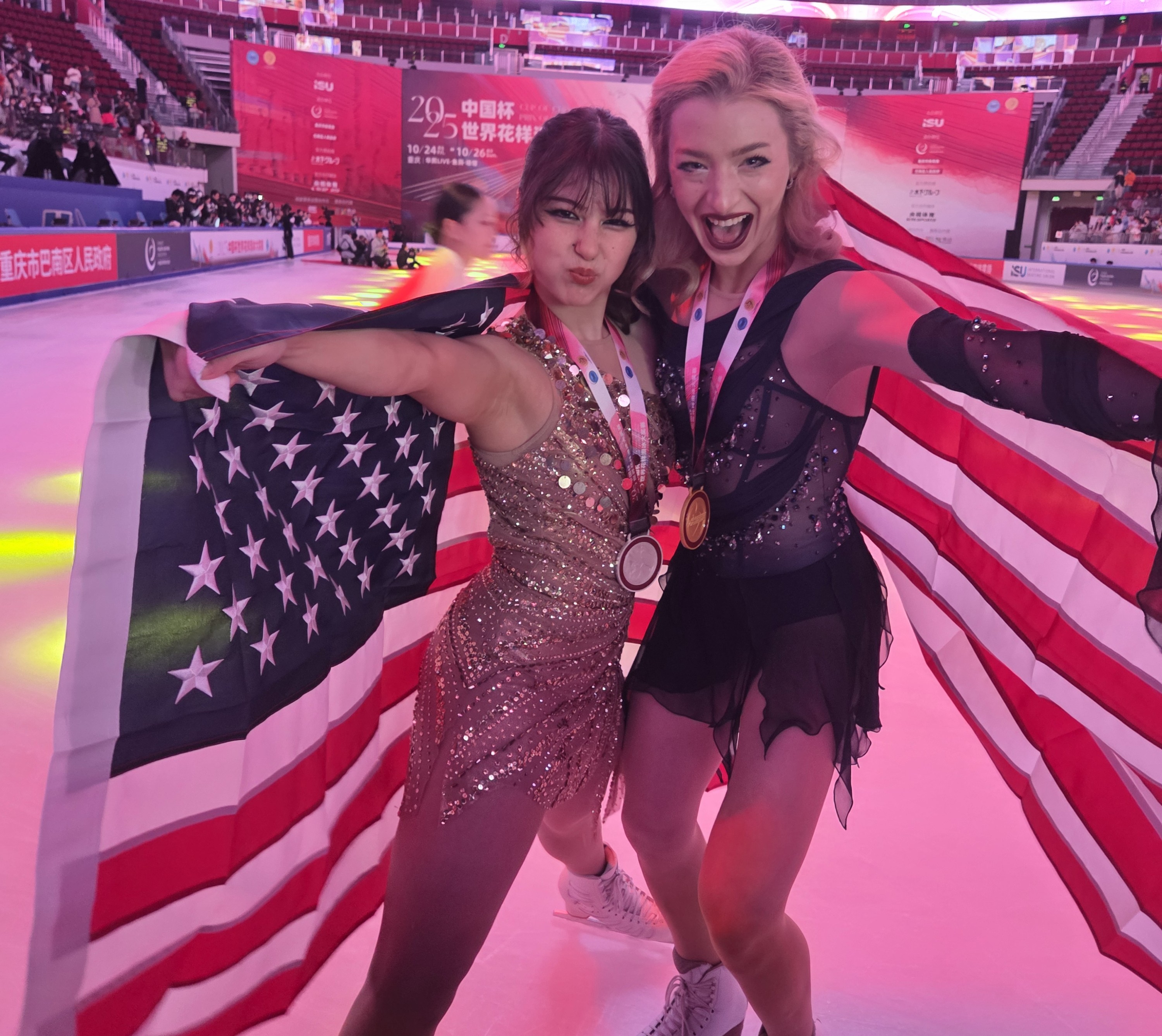 Alysa Liu (left) and Amber Glenn (right) strike a pose with silly looks on their faces while holding American flags behind them. Alysa (left) is a young woman with brown hair tied back in a bun and bangs wearing a gold sparkly dress. Amber is a young woman with blonde hair wearing a navy blue skating dress with jewel detailing. They both have 2025 Cup of China medals around their necks. 