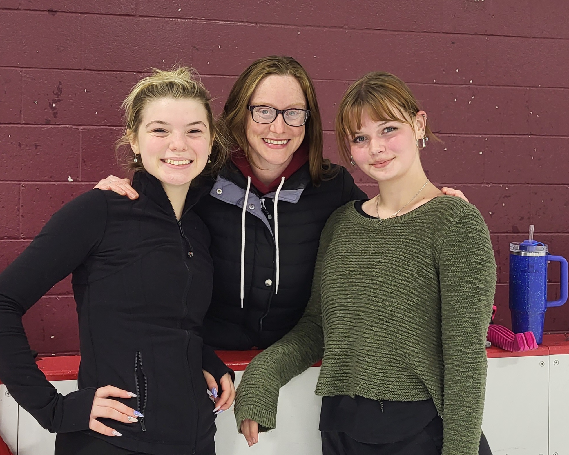 Carey Tinkelenberg (center) poses with two of her students. Carey is a white woman with shoulder length reddish hair and glasses. She holds her arms around two young female skaters. The one on the left is wearing a black jacket with blonde hair tied up in a bun, and the other is wearing a green sweater with brown hair tied back. 