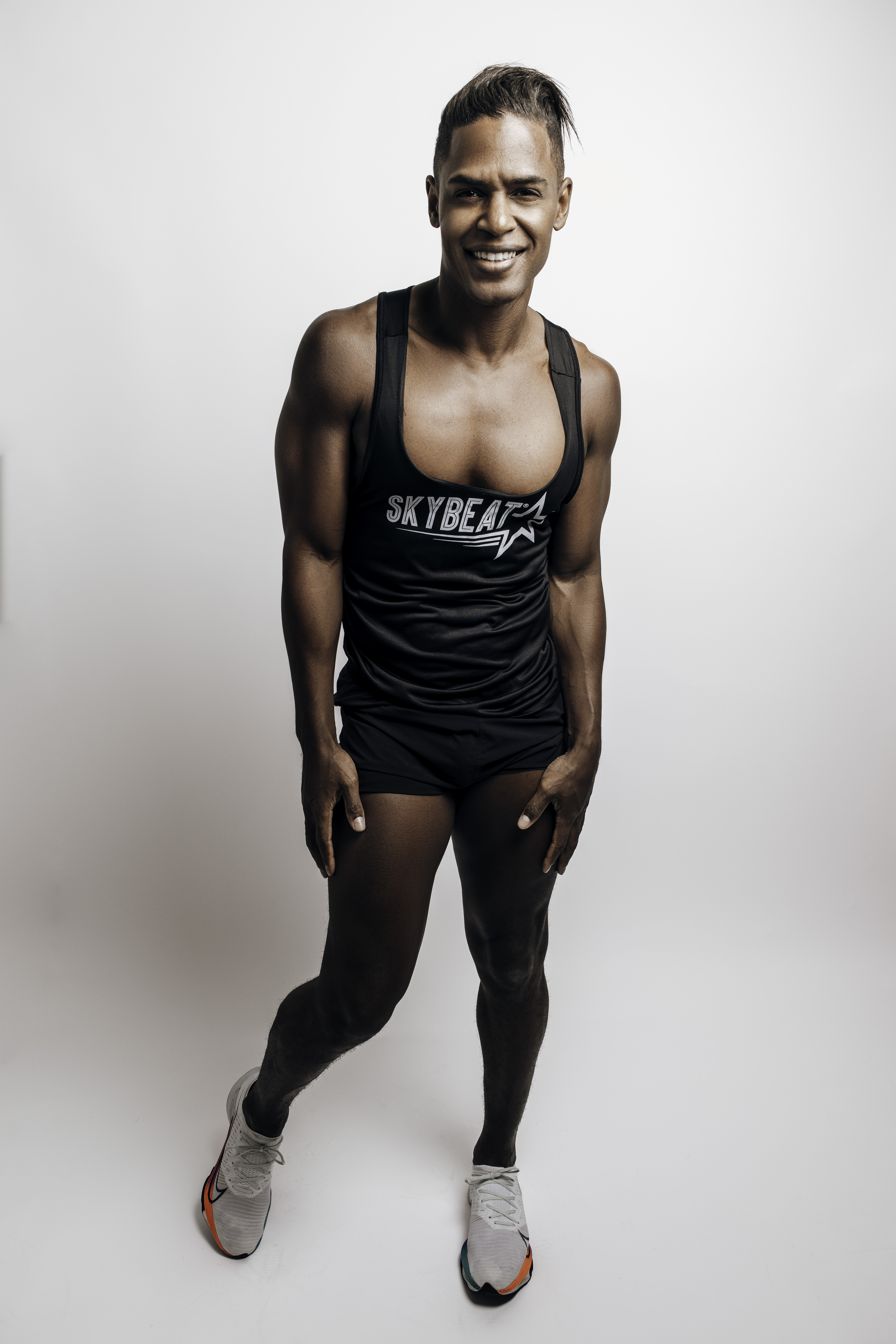 Skyler poses with his arms at his sides and his right knee slightly bent. He is a black man wearing a black tank top that says "SkyBeat" in white text with black shorts and white tennis shoes. He stands in front of a white background 