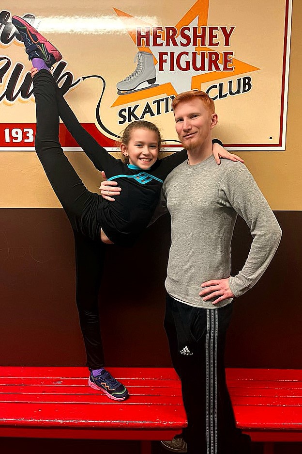 Rob and Alice Zinsmeister pose in front of a Hershey Figure Skating Club sign. Alice is a young girl wearing a log sleeve black shirt with a turquoise stripe across the front, black pants and grey and pink shoes. She stands holding one leg up in the air. Rob is a man with red hair wearing a long sleeve grey shirt. 