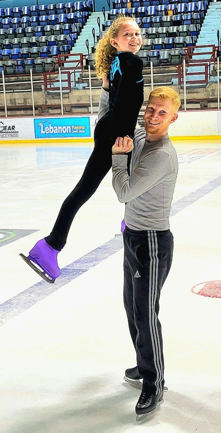 Rob holds Alice up on the ice. Alice is a young Alice is a young girl wearing a log sleeve black shirt with a turquoise stripe across the front, black pants and grey and pink shoes. Rob is a man with red hair wearing a long sleeve grey shirt. 