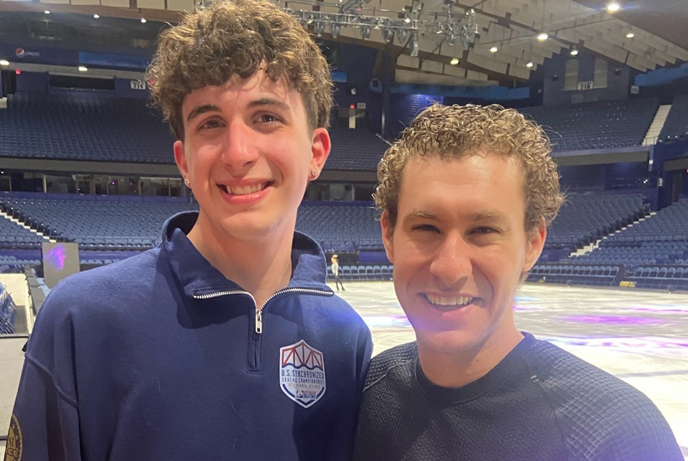 Quinn McCormick stands next to one of his skating idols, Jason Brown. Quinn is a young man with brown curly hair wearing a blue U.S. Synchronized Skating Championships quarter zip. Jason stands to his right. He is a slightly shorter man with brown curly hair wearing a black long sleeve shirt. 
