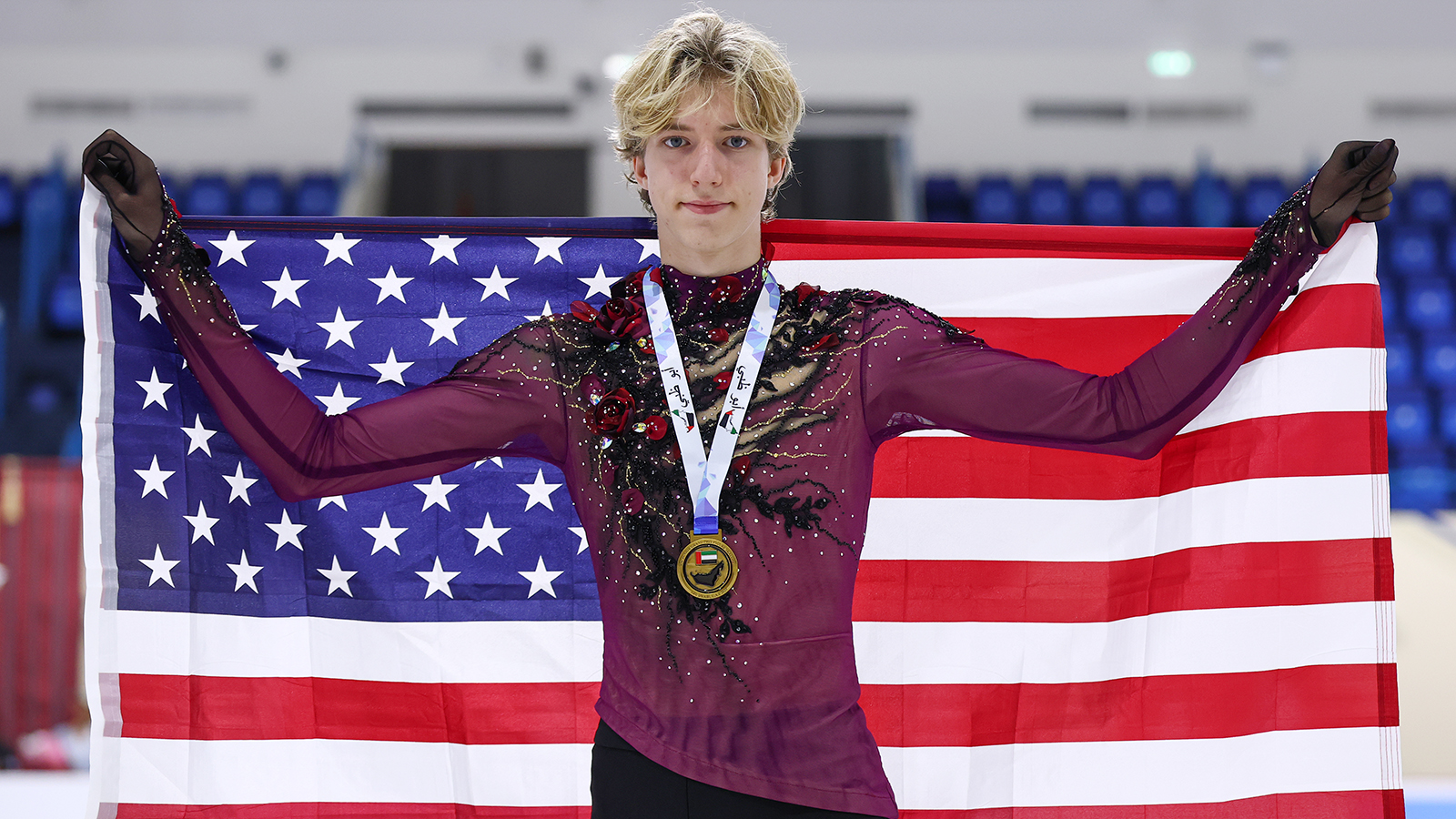 Photo taken from the waist up. Lucius Kazanecki stands holding an American flag behind him with a gold medal around his neck. Lucius is a tall young man with short blonde hair. He is wearing a long sleeve maroon top with black, gold and red floral detailing down the front and on the sleeves. 