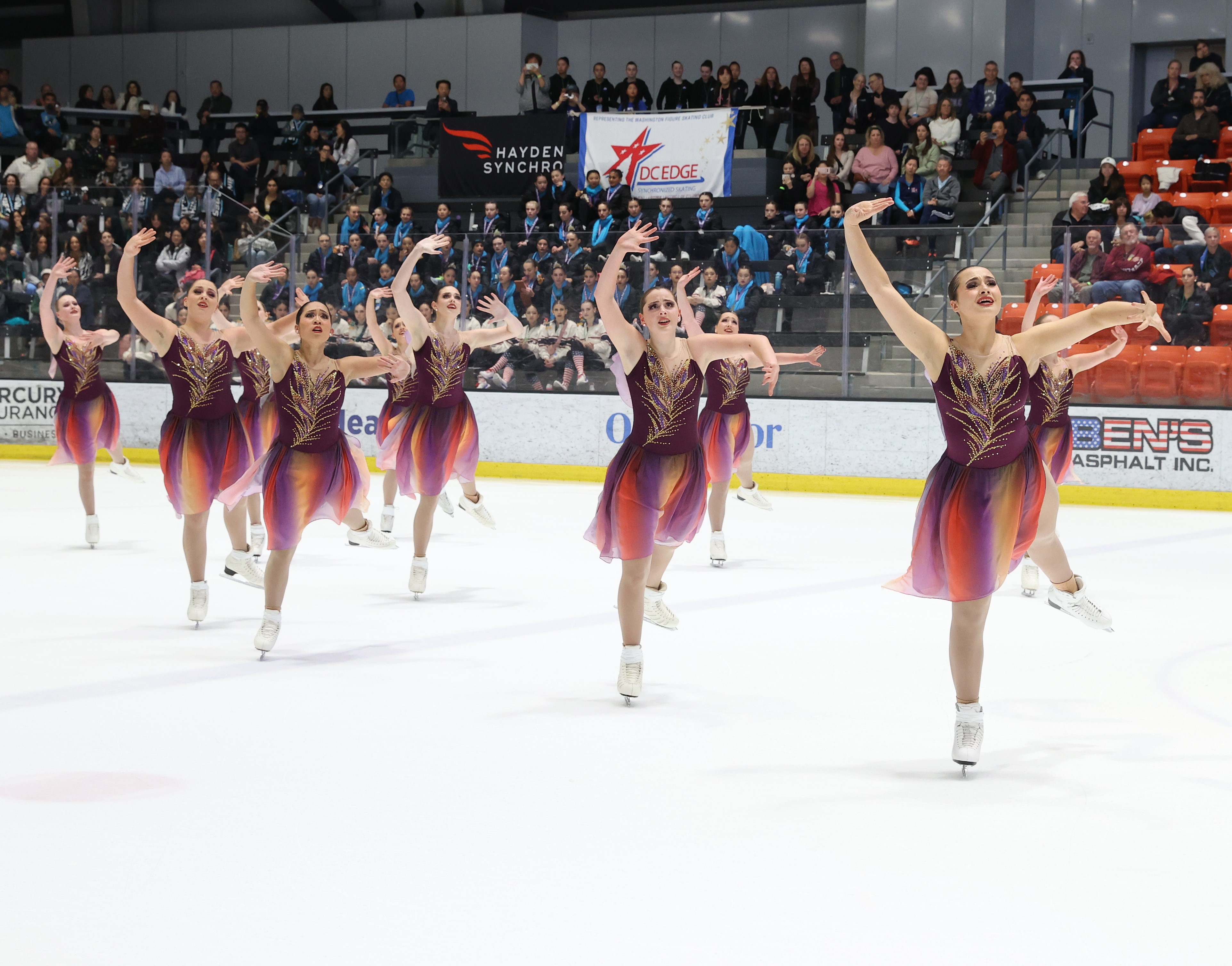 The Haydenettes compete in their short program wearing matching brown, yellow and purple skating costumes. They all skate in unison with their arms raised above their heads and with one leg extended behind them. 