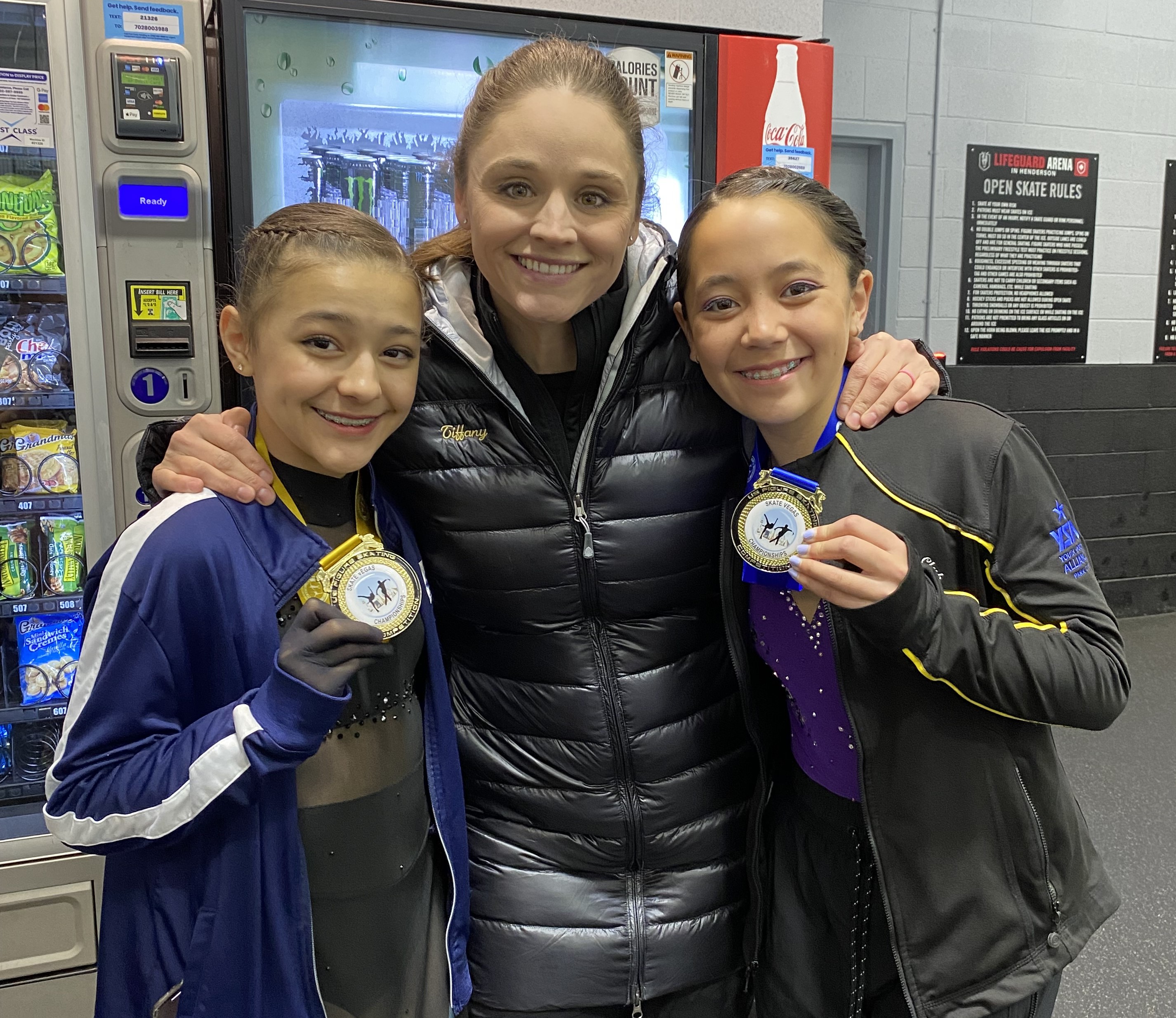Tiffany McNeil (center) poses with two of her skaters. Tiffany (center) is an older white woman wearing her hair back in a ponytail and a black winter jacket. She stands in between two young female skaters showing off their medals they won in competition 