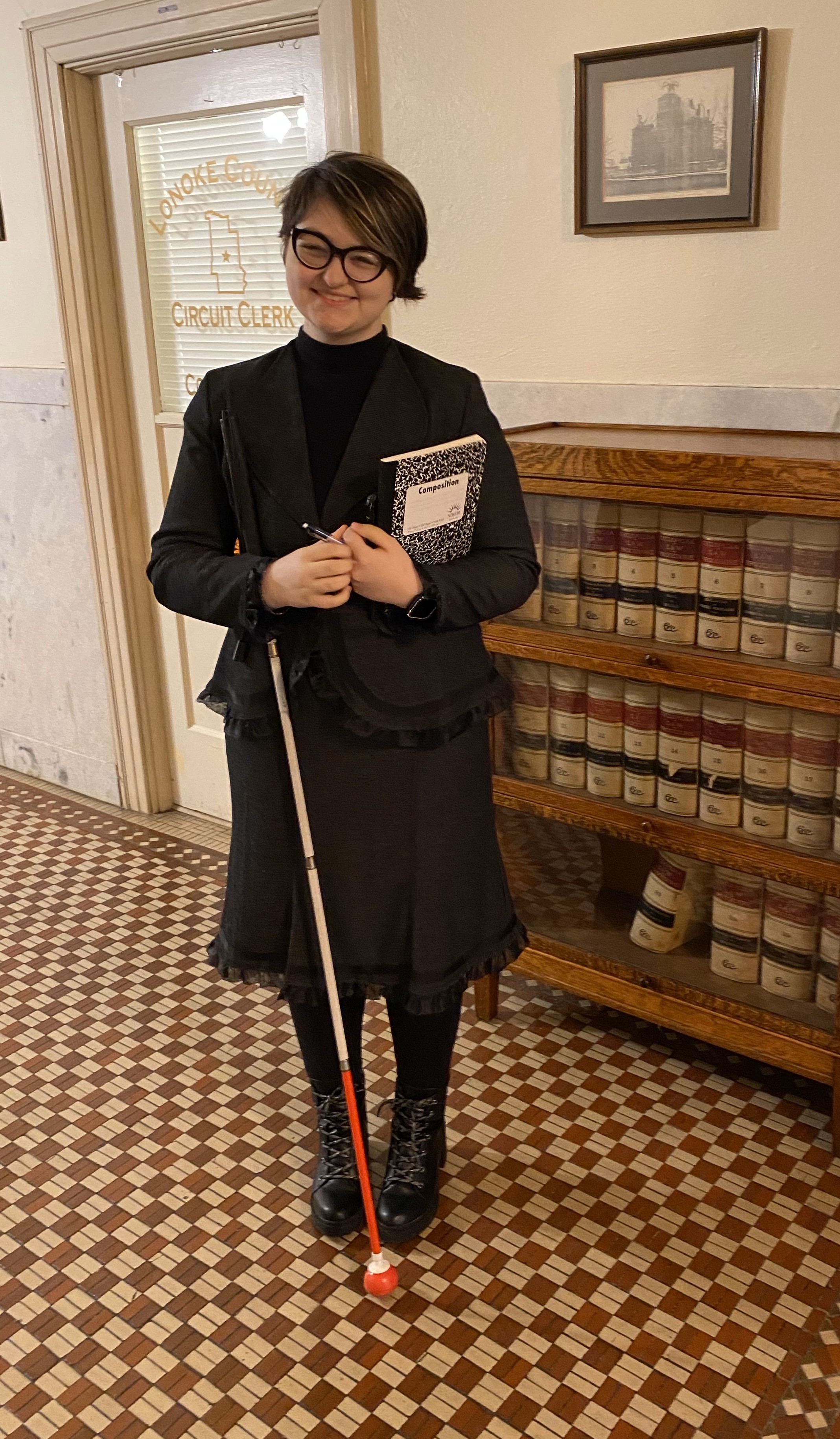 Tobi Ellis smiles at the camera holding a notebook in one hand and resting their mobility cane on their other arm. They have short blonde hair and thick rimmed glasses. They are wearing a black turtleneck with a black blazer on top, a black skirt, black tights and black combat boots. They stand in front of a bookshelf and a door that says "Lonoke County Circuit Clerk" in gold lettering. 