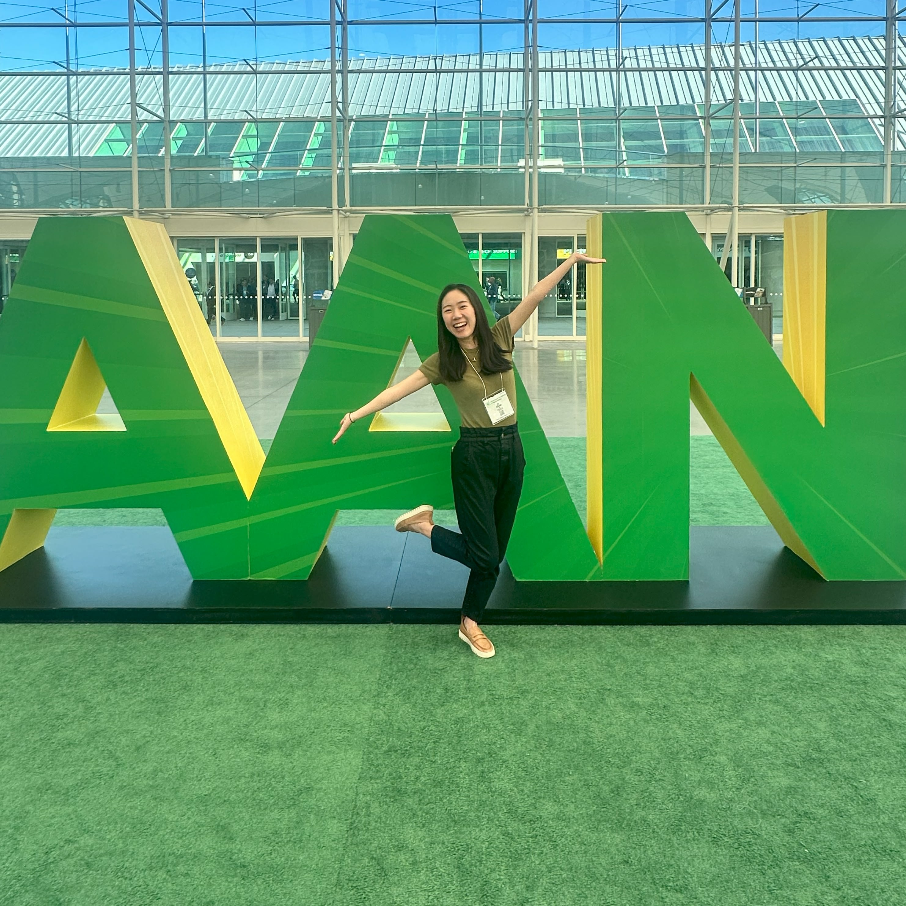 Ai Ohno stands in front of a big green and yellow sign with the letters "AAN". She stands in front, smiling with her arms raised. Ai is a young Asian woman with long black hair wearing a green tshirt and black pants. 