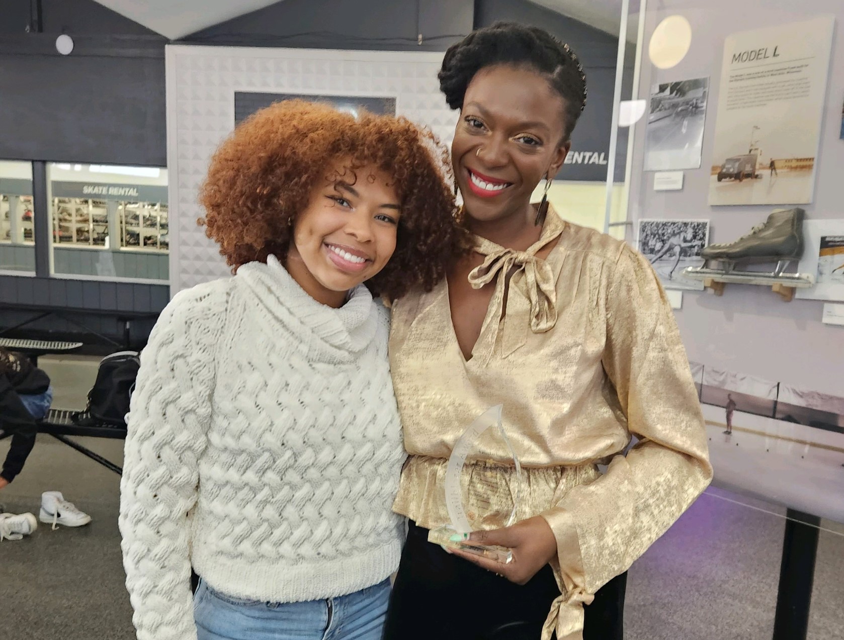 Kadari Taylor-Watson (R) poses with skater Starr Andrews (L). Andrews is a young Black woman with an afro. She is wearing a knit white sweater and jeans. Taylor-Watson is a Black woman wearing a gold top with a bow tied at the neck and black pants. Taylor-Waton holds a glass tear drop shaped award 