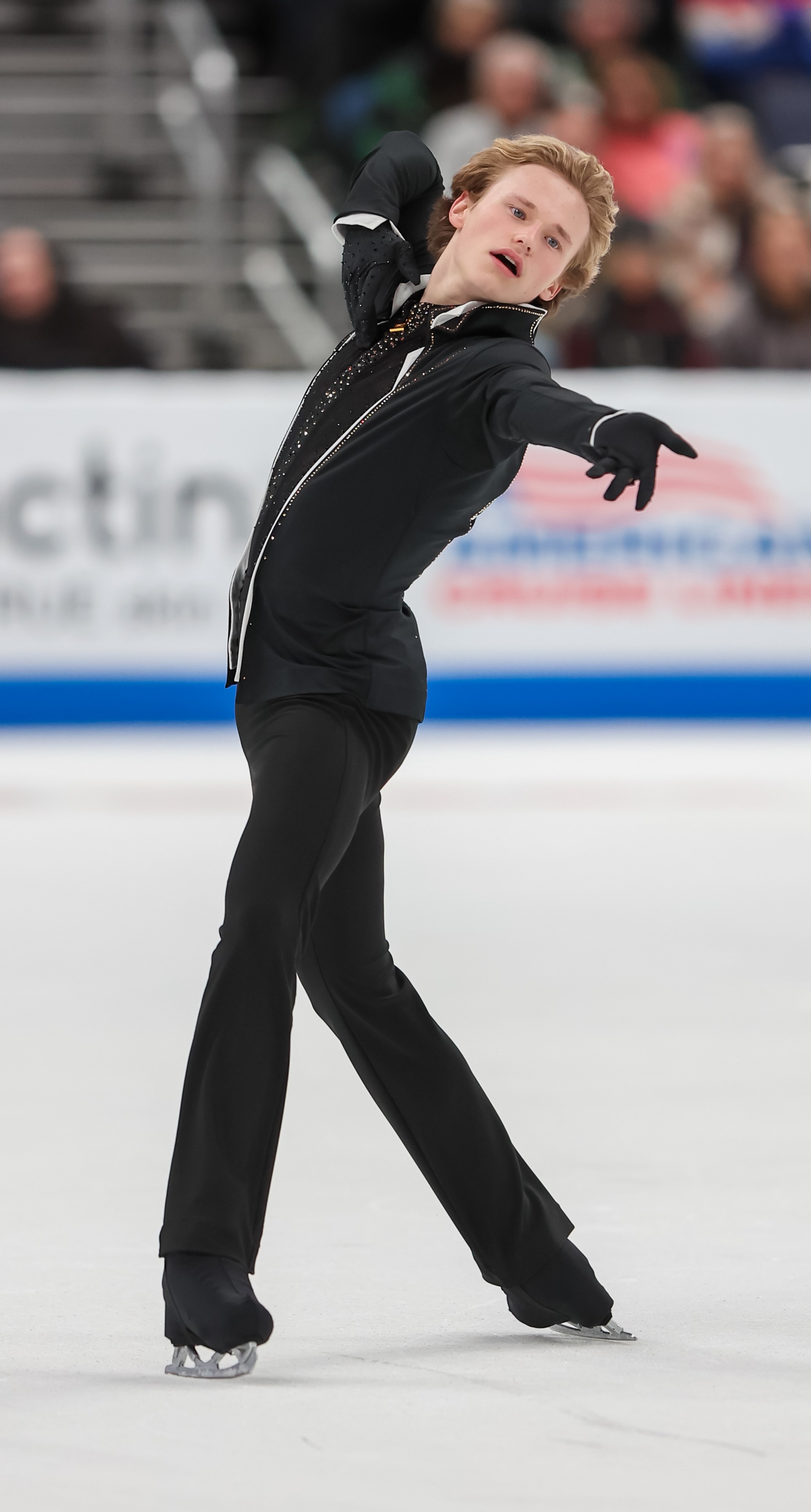 Ilia Malinin strikes a pose with one arm extended out in front of him. He is wearing a black top with white lining and a black tie and black pants.