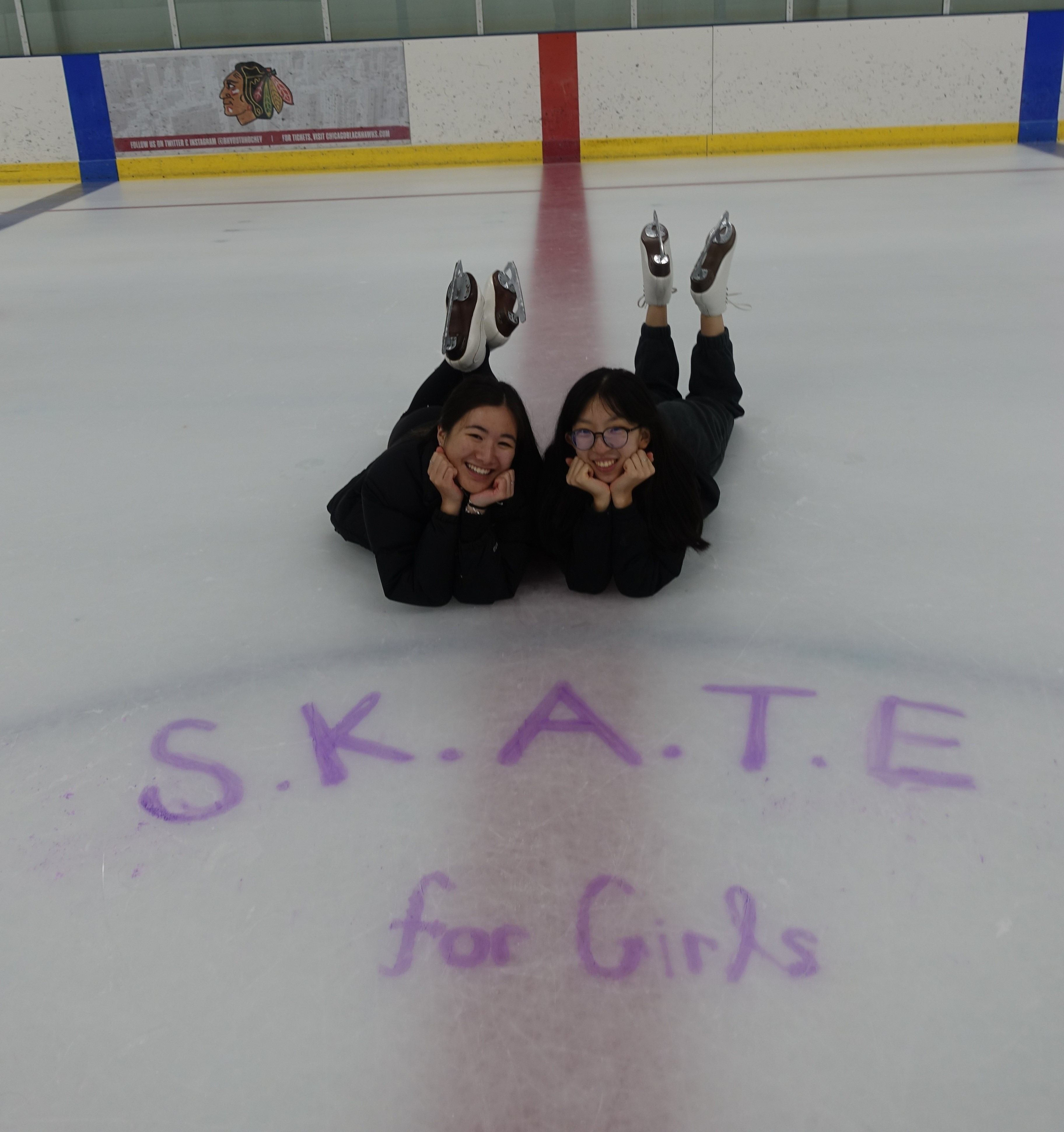 Mayumi and frend Jasmine Fang lay on their stomachs on the ice. S.K.A.T.E for Girls is written on the ice. Mayumi (left) is a young Asian woman with long black hair. She is wearing a black long sleeve shirt and black pants. Jasmine (right) is a young Asian woman with long black hair and glasses. She is also wearing a black long sleeve shirt and black pants 