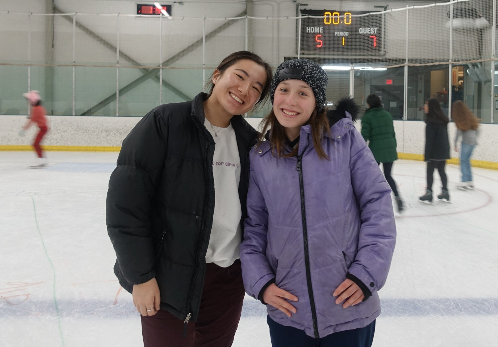 Mayumi smiles with one of her students. Mayumi (left) is a young Asian woman with long black hair tied back in a ponytail. Mayumi (left) is wearing a white S.K.A.T.E. for Girls sweatshirt woth a black jackt and maroon pants. Her student (right) is a young white woman with shoulder length brown hair under a black and white beanie. She is wearing a lavender colored jacket and blue pants