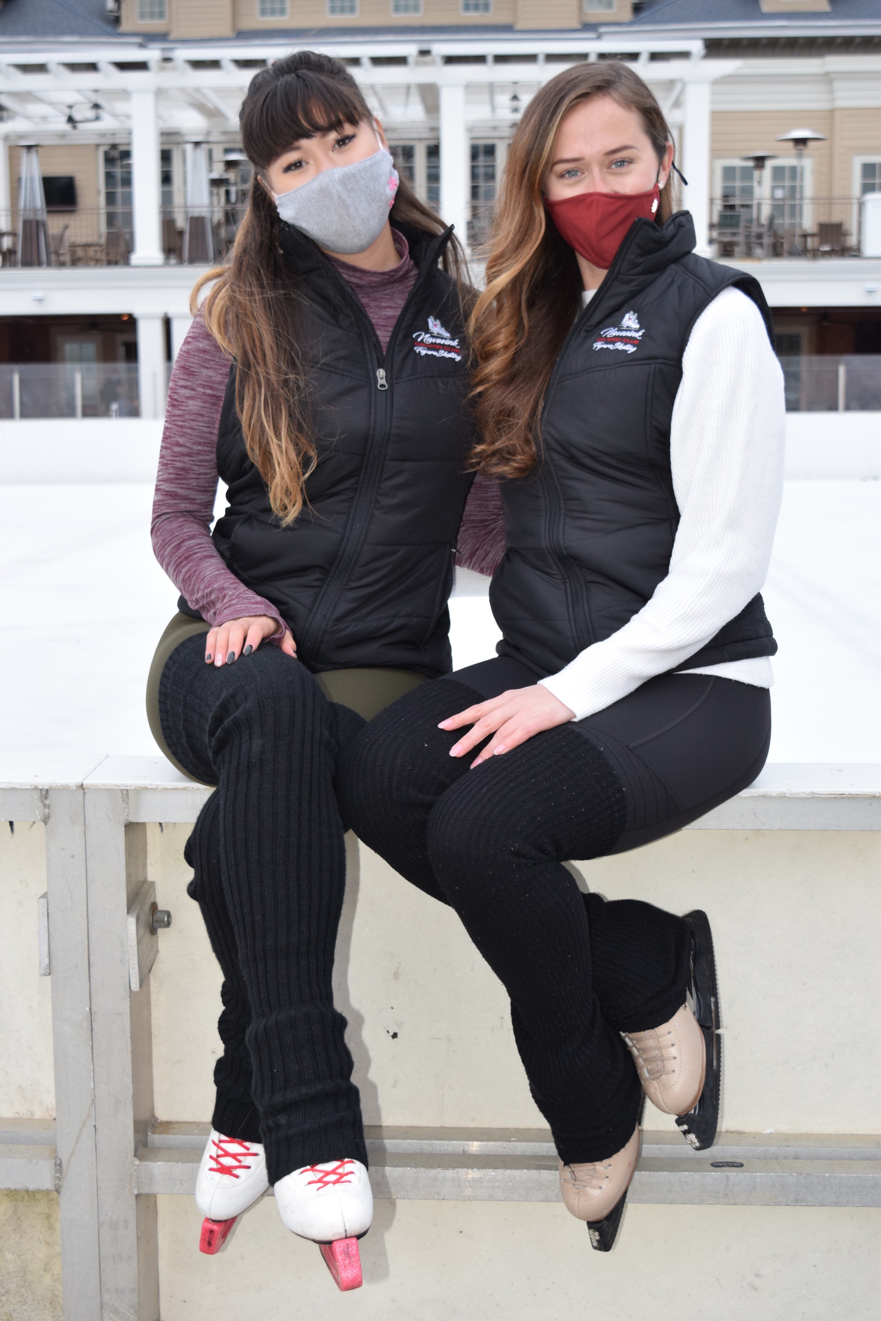 Coaches Mariko Heimbach and Jennifer Gruver sit side by side wearing masks and skates.