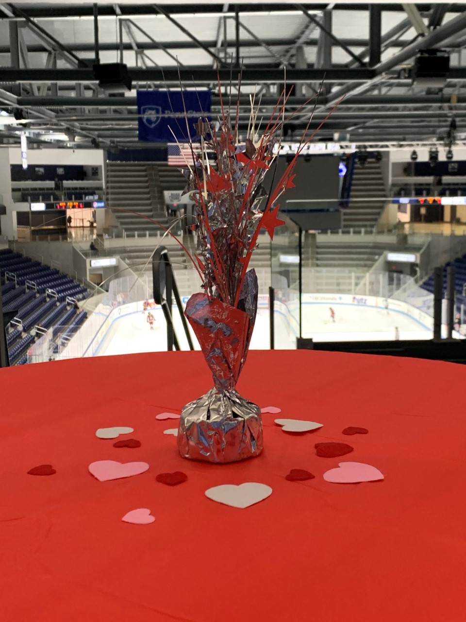 A red valentines day themed centerpiece with hearts sits on a table with a red table cloth overlooking an ice rink. 