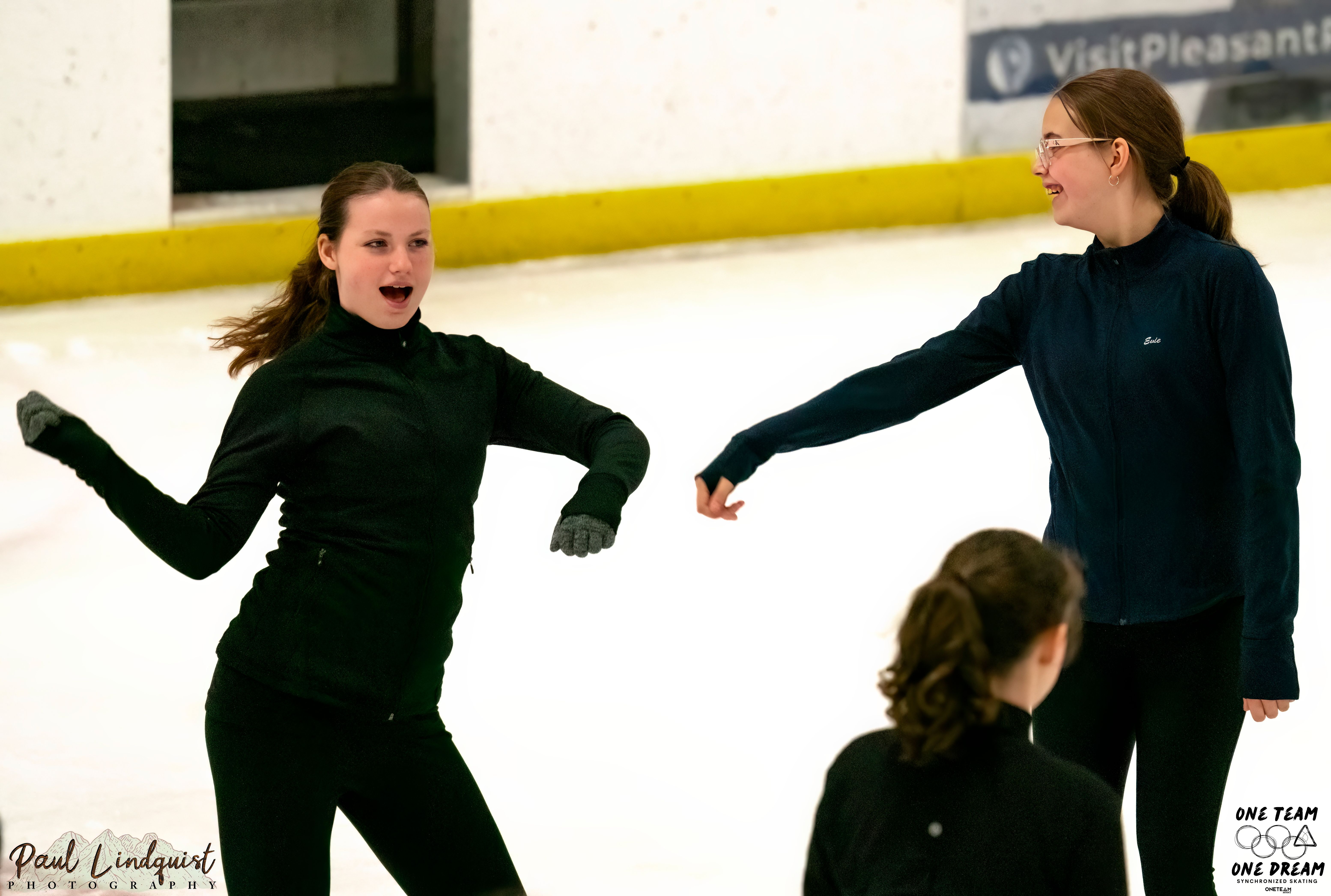 Two young girls goof off on the ice. One girl (left) looks like she is dancing. She has long brown hair tied back in a ponytail and is wearing all black. The other skater (right) smiles and laughs looking at her friend. She also has long brown hair in a ponytail and has glasses and braces. 