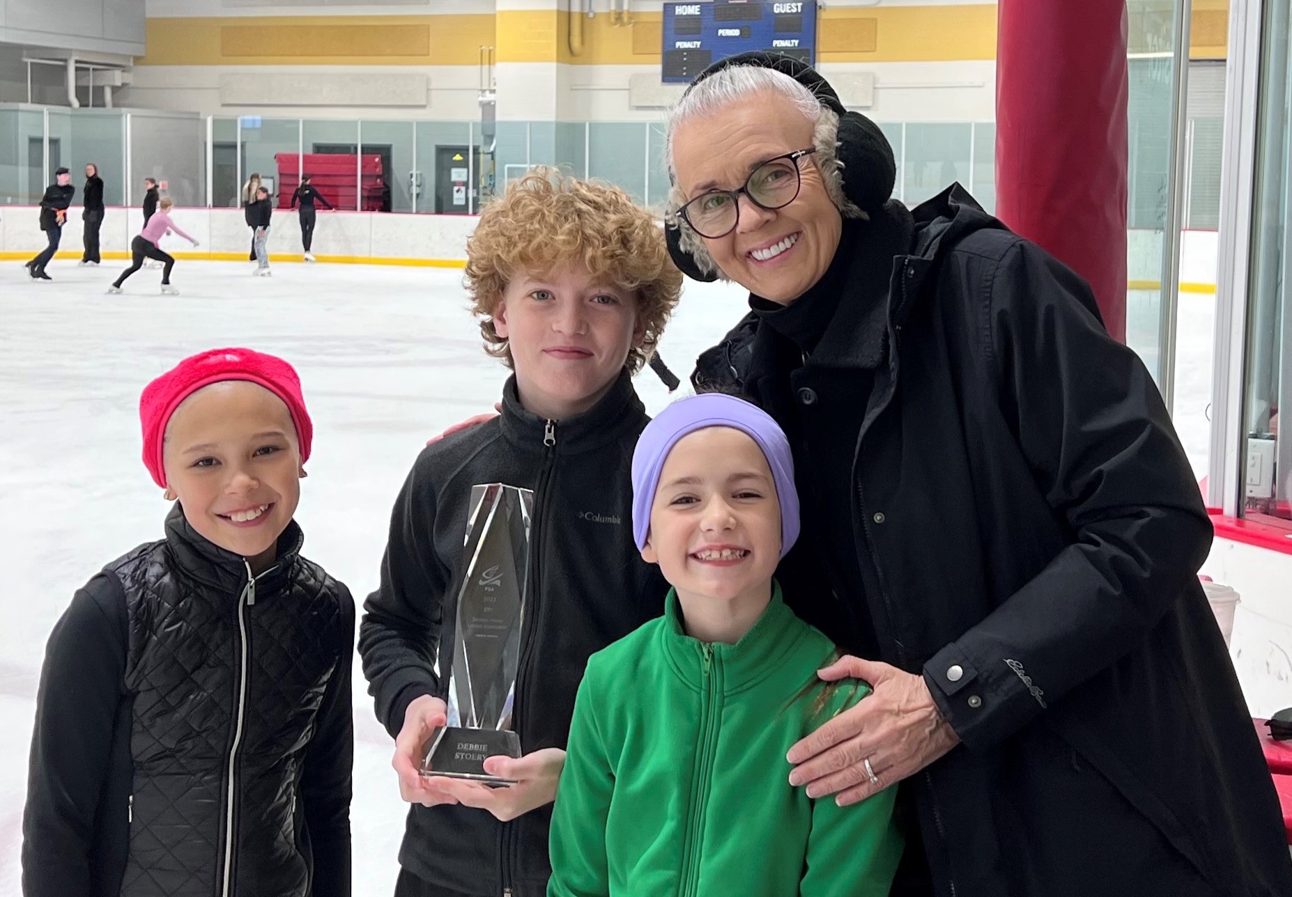 Debbie Stoery poses with three young skaters on the ice. Debbie is an older white woman with white hair tied back. She has on glasses white ea muffs. She poses with two young female skaters and one young male skaters all wearing winter jackets. 