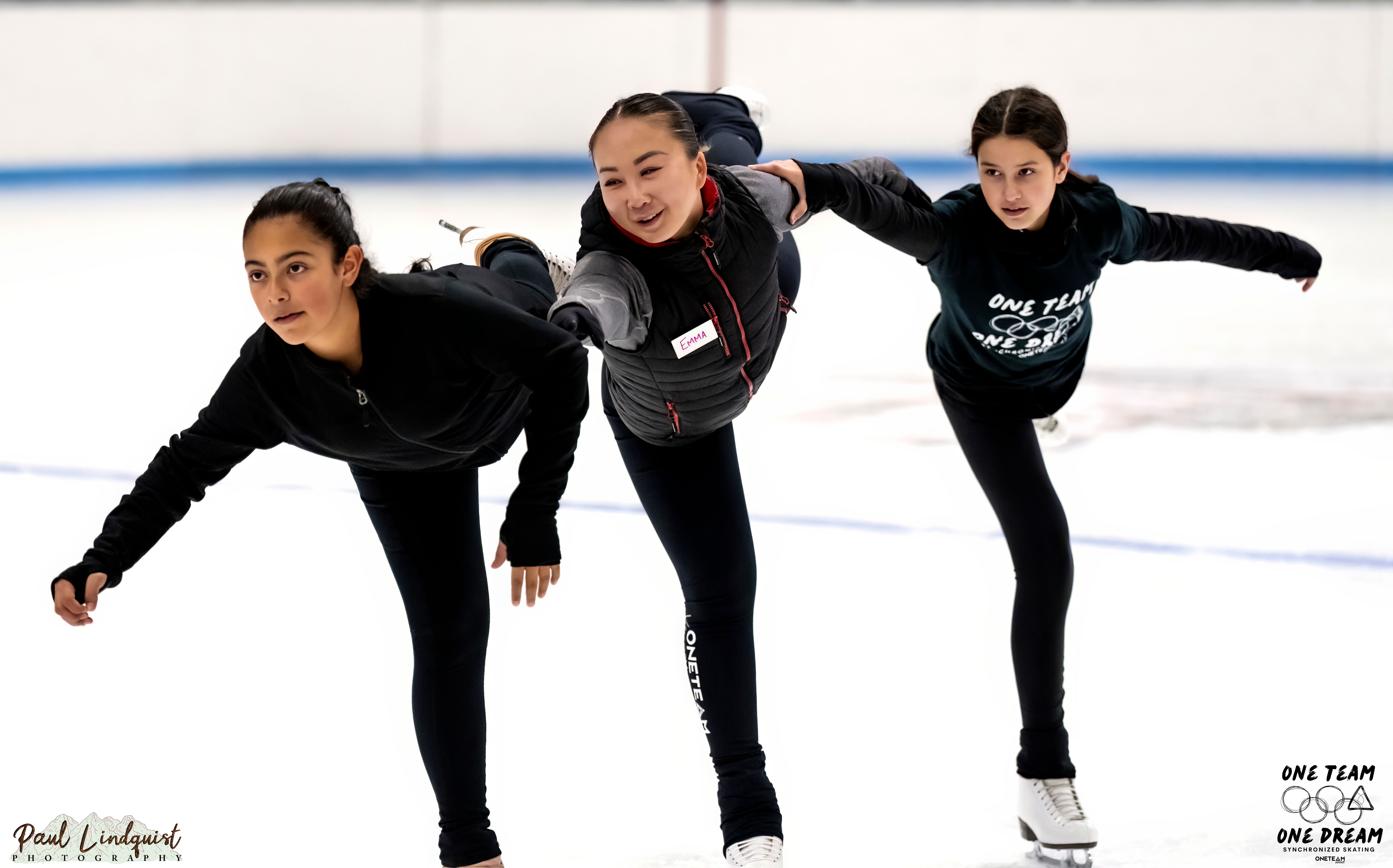 Three skaters hold on to each other while they skate with one leg extended behind them. The skaters are three young girls wearing all black. 