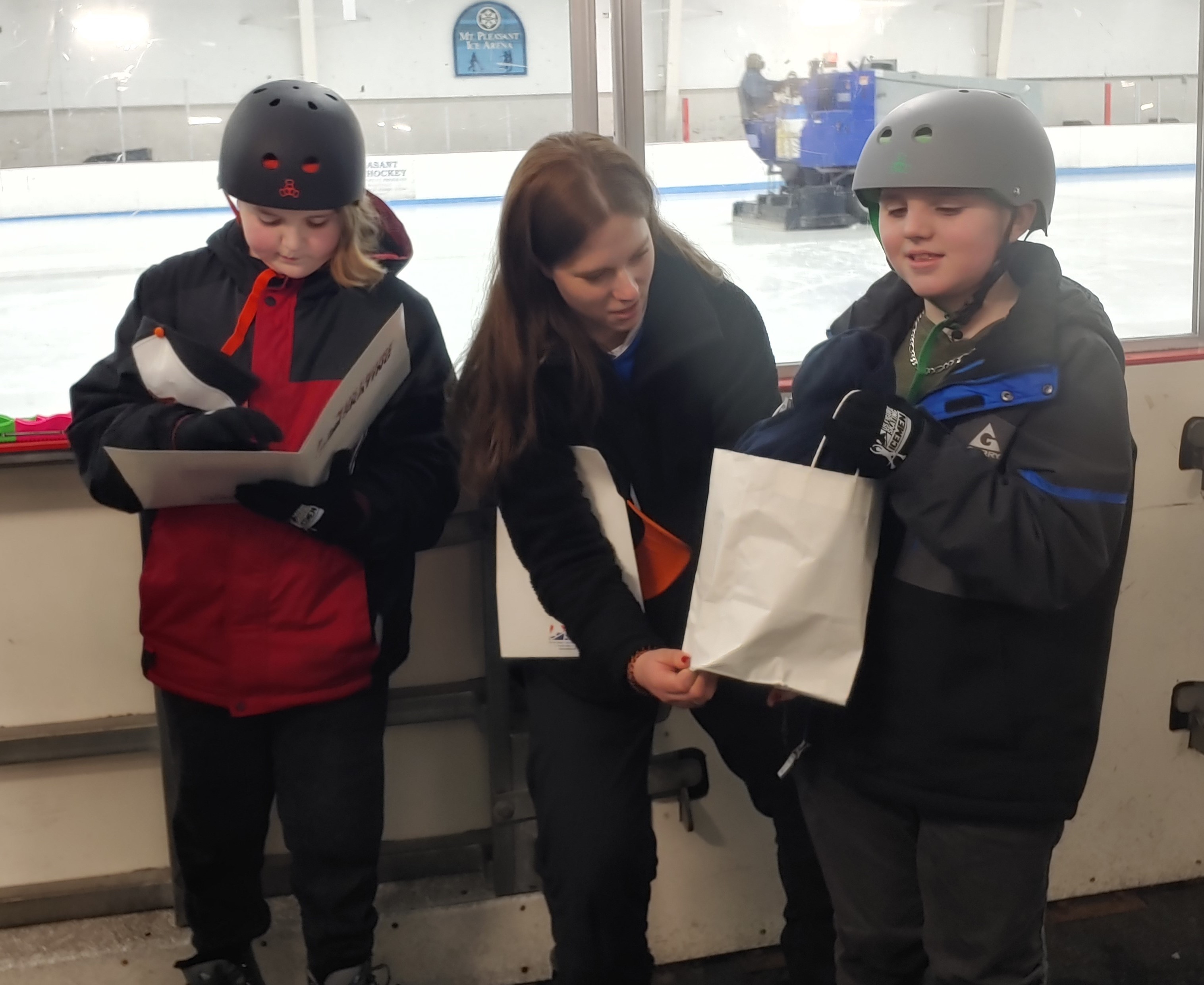 Noah (right) is given a white gift bag with a navy blue tshirt inside with the name of the club on the front. Noah is a young boy wearing a grey helmet and a blue and grey jacket. His brother Logan stands on the left side of the photo wearing a black helmet and red jacket. He looks down at a white folder with the U.S. Figure Skating logo on the front