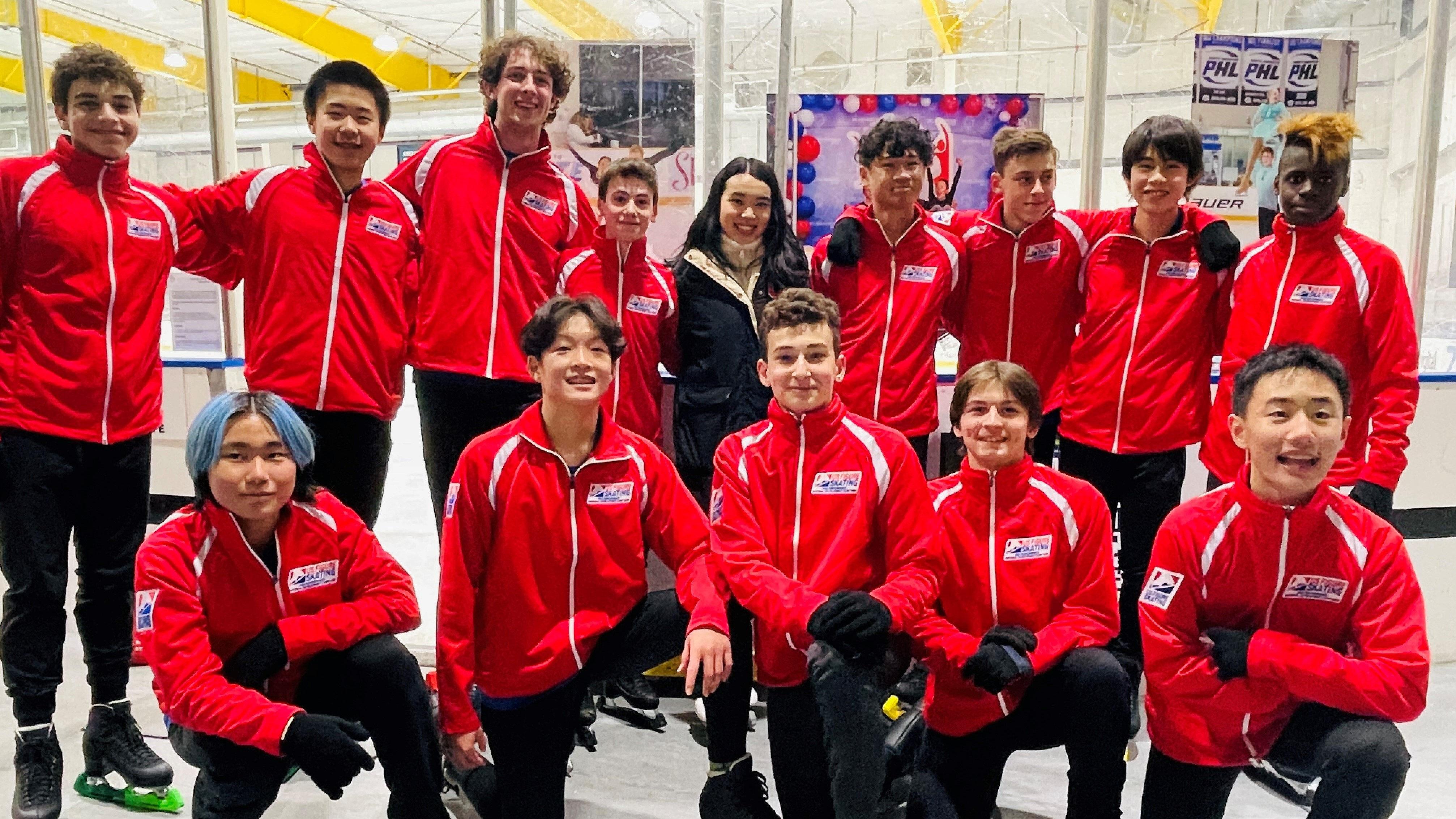 Marlo Rosen along with a group of men's skaters pose with Team USA's Karen Chen. Marlo is a young boy with curly brown hair. Karen stands in the middle of the group weating a black winter jacket. She is an Asian woman with long black hair. All of the young skaters are wearing their red National Development Team jackets. 
