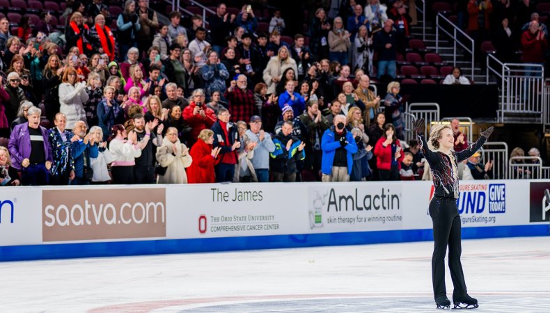 Ilia Malinin salutes the crowd with his arms outstretched and a smile on his face as people in the stands cheer. Ilia is a young man with short blonde hair wearing a black top with red and white stripes and black pants. 