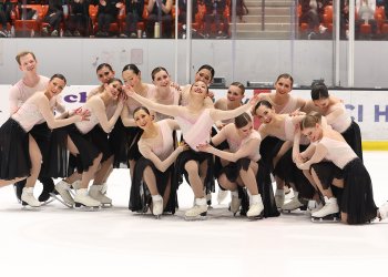 The Haydenettes strike a beginning pose, leaning on each other. The skater in the middle looks up with a smile on her face and her arms outstretched. They are all wearing matching skating costumes with a light pink long sleeve top and black skirt. One male skater on the far left is wearing a matching pink top with black pants.
