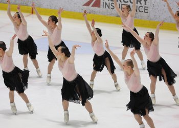 The Haydenettes skate in unison in a group with their hands raised above their heads. They are all wearing matching costumes with a long sleeve pink top and a black skirt.