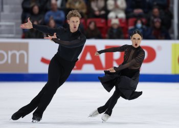 Madison Chock and Evan Bates skate side by side with both athletes pointing their arms to the left in unison. Evan (left) is a tall man with short blonde hair wearing a long sleeve black top and black pants. Madison (right) is a woman with black hair tied back in a bun. She is wearing a black skating costume with long sleeves and a long black skirt