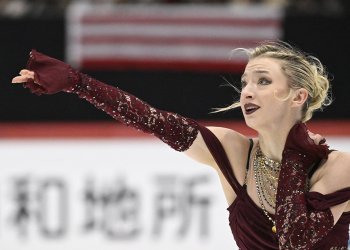 Photo taken from the waist up of Amber Glenn. She looks off to the left with one arm outstretched. Amber is a young woman with blonde hair tied back in a bun wearing a maroon skating costume with chunky gold necklaces and matching maroon gloves.