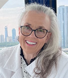 Headshot of Debbie Stoery. Debbie is an older woman with long white hair wearing glasses and a white jacket