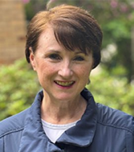 Headshot of Gloria Leous. Gloria is an older woman with short red hair wearing a navy blue jacket